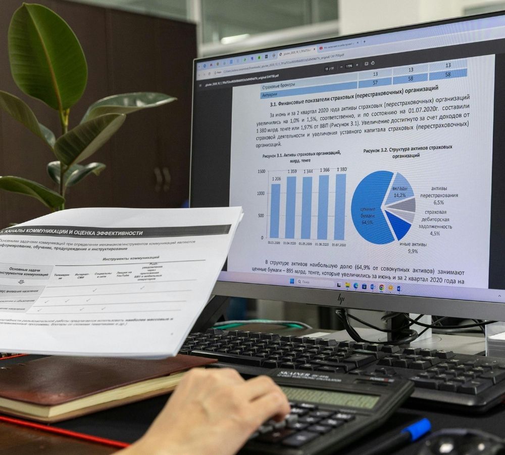 A person using a computer views financial charts and text on the screen while holding a printout