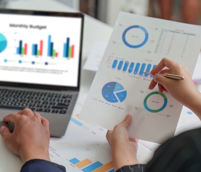 Hands analyzing financial charts on paper and a laptop screen displaying a monthly budget with colorful graphs