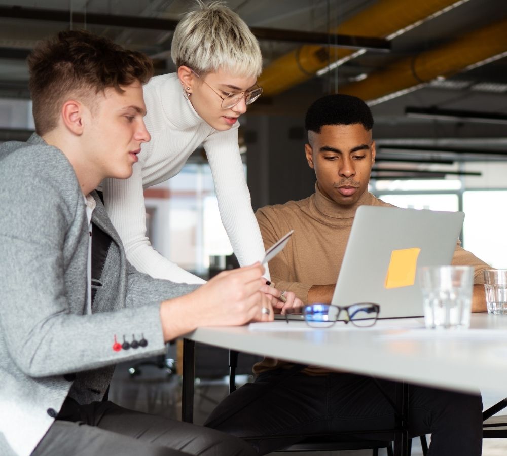 Three colleagues collaborate at a table; two men, and one woman leaning in