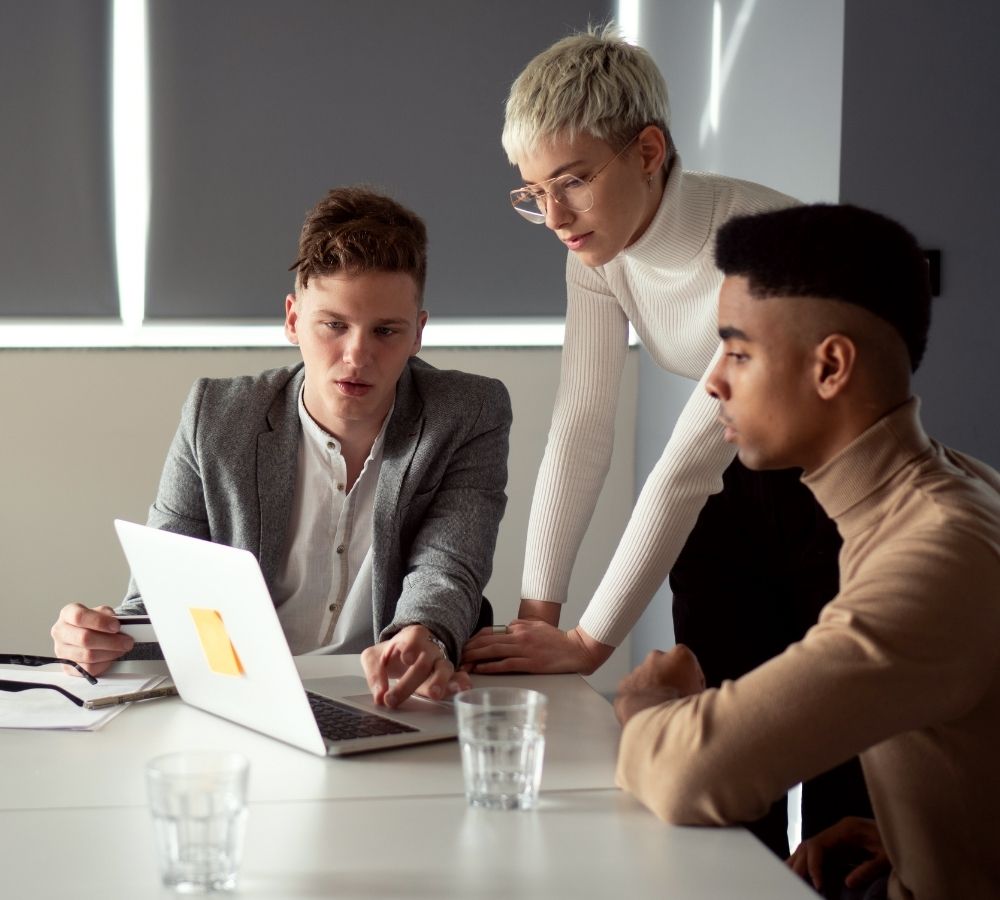 Three people in a modern office collaborate at a table, focused intently on a laptop