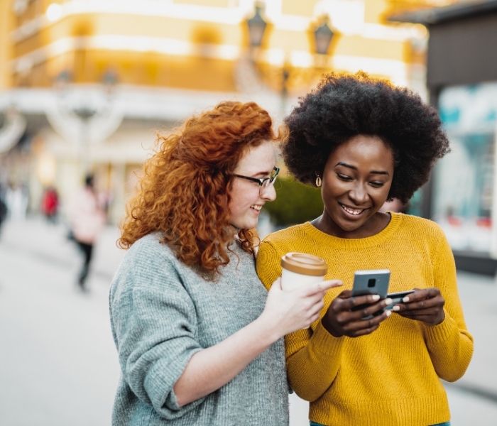 Two women smile while looking at a phone. One holds a coffee cup