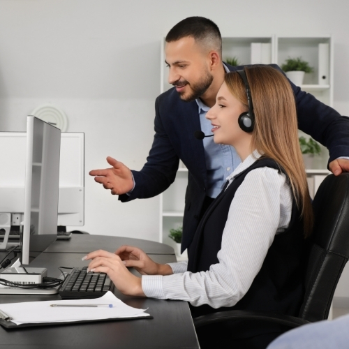 A woman wearing a headset sits at a desk while a man beside her gestures at a computer screen